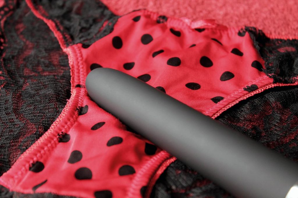 Ein schwarzer klassischer Vibrator liegt auf ein rotes Damen-Höschen, welches schwarz gepunktet ist.